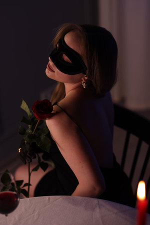 Elegant woman in black eye mask with rose indoors in eveningの写真素材