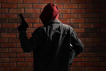 Thief in hoodie with gun against red brick wall, back viewの写真素材