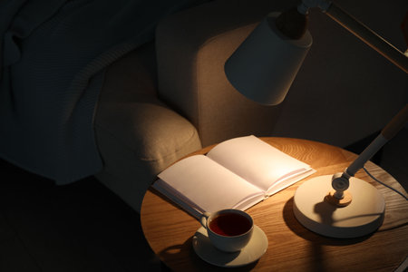 Stylish lamp, book and cup of tea on side table near sofa indoors