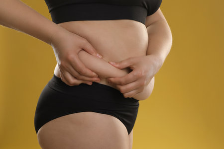 Woman touching belly fat on goldenrod background, closeup. Overweight problemの写真素材