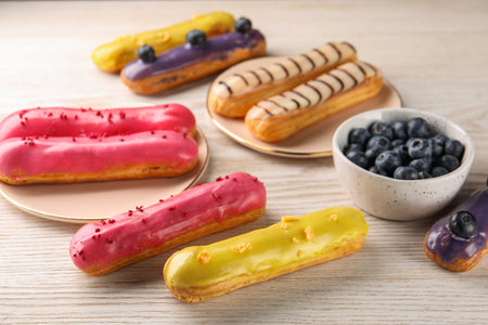 Different tasty glazed eclairs and blueberries on light wooden table, closeup
