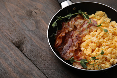 Delicious scrambled eggs with bacon in frying pan on wooden table. Space for textの写真素材