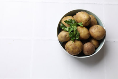 Tasty young boiled potatoes with parsley in bowl on white tiled table, top view. Space for textの写真素材