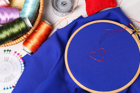 Blue cloth with hoop, embroidered heart, thread and sewing needle on light table with tools, flat layの写真素材