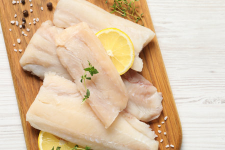 Pieces of raw cod fish, lemon and spices on white wooden table, top view