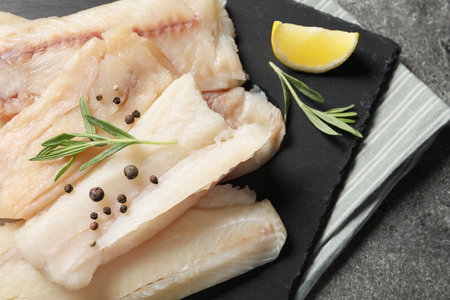 Pieces of raw cod fish, rosemary, peppercorns and lemon on grey table, closeup