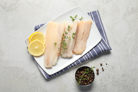 Pieces of raw cod fish, peppercorns and lemon on light gray table, top view
