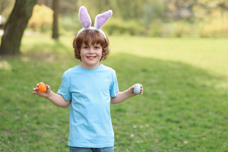 Easter celebration. Cute little boy in bunny ears holding painted eggs outdoors, space for textの写真素材