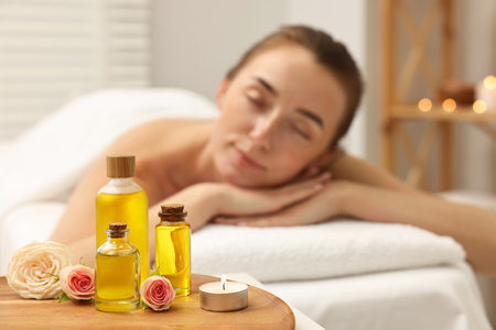 Aromatherapy. Woman relaxing on massage couch in spa salon, focus on bottles of essential oils, burning candle and rose flowersの写真素材