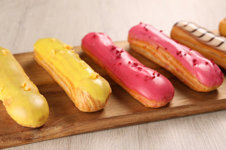 Board with different tasty glazed eclairs on light wooden table, closeup