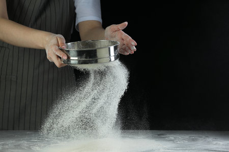 Woman sieving flour at table against black background, closeup. Space for text