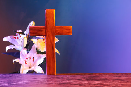 Wooden cross and beautiful lily flowers on table in color lights, space for text. Religion of...