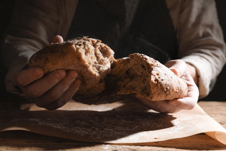 Man breaking loaf of fresh bread at wooden table, closeup