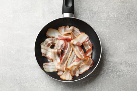 Delicious bacon slices in frying pan on light gray table, top view