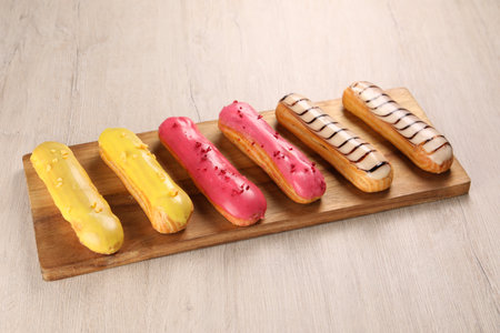 Board with different tasty glazed eclairs on light wooden table