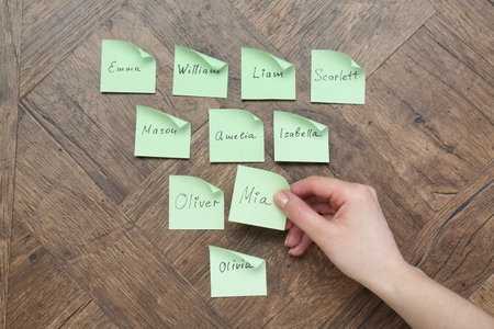 Woman holding paper sticker with name at wooden table, top view. Choosing baby's name