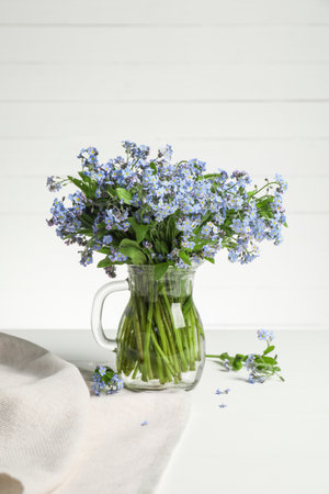 Bouquet of beautiful forget-me-not flowers in glass jug and blue cloth on white tableの写真素材