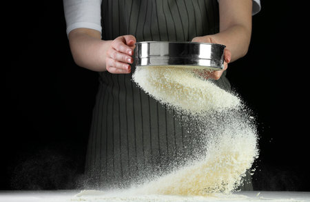 Woman sieving flour at table against black background, closeup