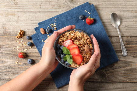 Woman holding bowl of tasty granola with berries, nuts and mint at wooden table, top viewの写真素材