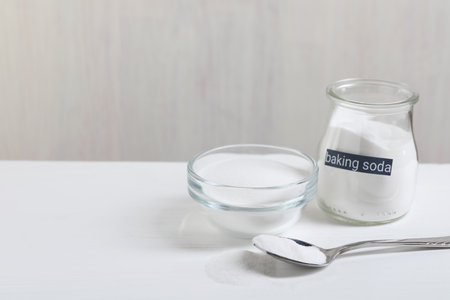 Baking soda in bowl, glass jar and spoon on white wooden table, space for text