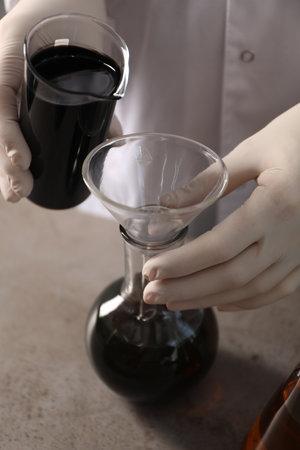 Woman pouring black crude oil from beaker into flask at gray table, closeup