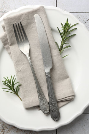 Stylish setting with cutlery, napkin, rosemary and plate on light tiled table, top view