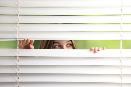 Young woman looking through window blinds on blurred backgroundの写真素材