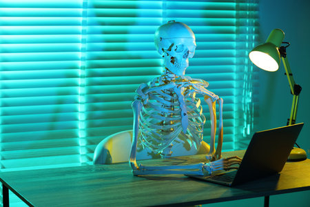 Waiting concept. Human skeleton sitting at wooden table with laptop indoors