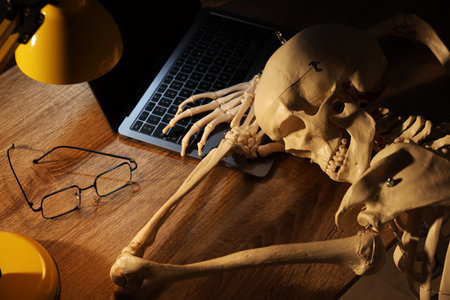 Waiting concept. Human skeleton sleeping at wooden table with laptop