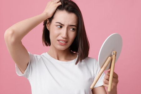Sad woman with hair loss problem looking at mirror on pink backgroundの写真素材