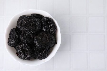 Tasty dried plums (prunes) in bowl on white tiled table, top view. Space for textの写真素材