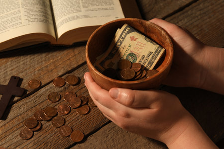 Donate and give concept. Woman holding bowl with coins and dollar banknotes at wooden table, closeup