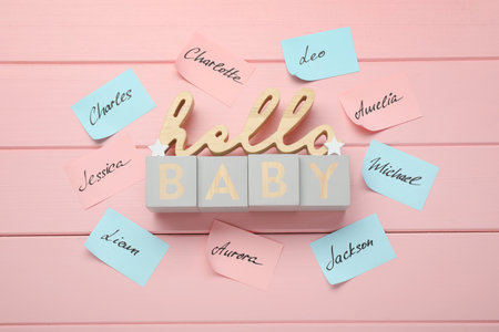 Cubes with phrase Hello Baby and paper stickers with different names on pink wooden background, flat...