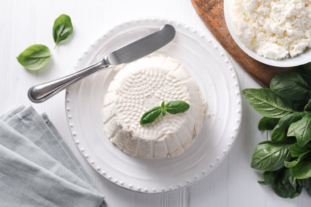 Fresh ricotta (cream cheese) with basil and knife on white wooden table, flat layの写真素材