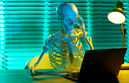 Waiting concept. Human skeleton sitting at wooden table with laptop indoors
