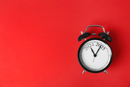 Black alarm clock with hands pointing at numbers 2024 and 2025 on red background, top view....