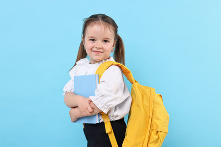 Cute little girl with book and backpack on light blue background