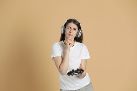 Thoughtful woman in headphones with controller on beige background