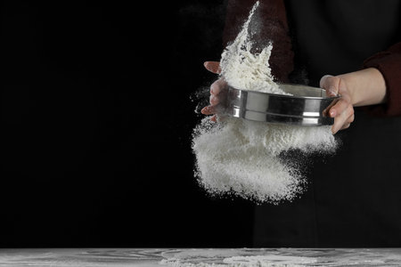 Woman sieving flour at table against black background, closeup