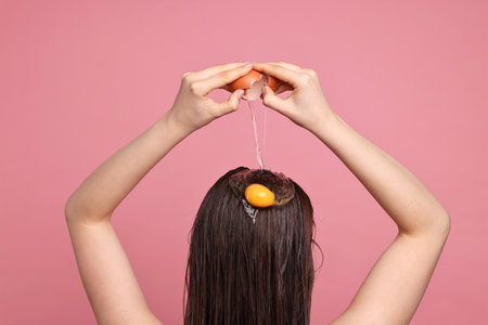 Woman breaking egg onto her head against pink background, back view. Natural hair maskの写真素材