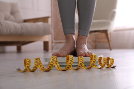 Eating disorder. Woman standing on floor scale and measuring tape indoors, closeupの写真素材