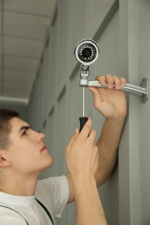 Technician with screwdriver installing CCTV camera on wall indoors