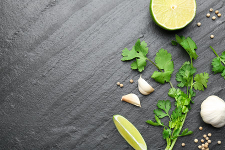 Fresh coriander leaves, dried seeds, garlic and lime on black textured table, flat lay. Space for...