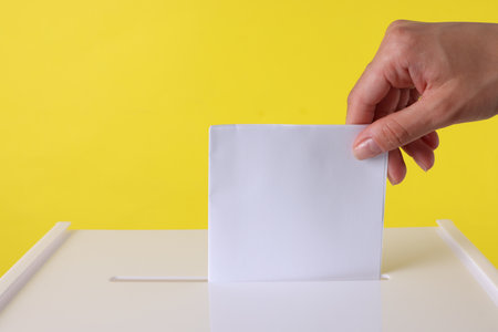 Woman putting her vote into ballot box against yellow background, closeupの写真素材