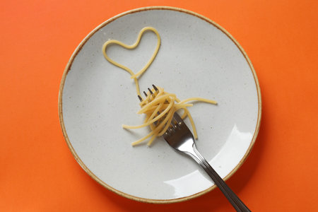 Heart made of tasty spaghetti and fork on orange background, top view