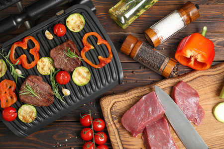 Electric grill with meat, spices, knife and vegetables on wooden table, flat layの写真素材