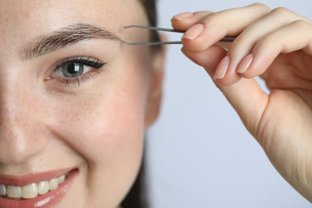 Young woman plucking eyebrow with tweezers on light background, closeup