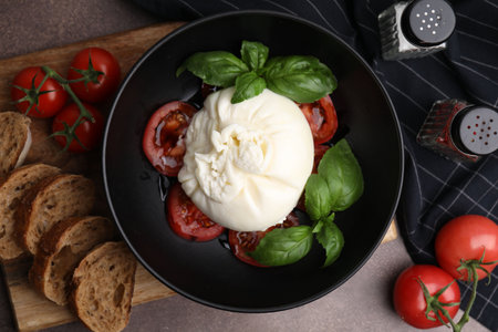 Delicious burrata cheese, tomatoes, basil and bread in bowl on brown table, flat layの写真素材