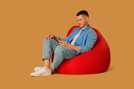 Handsome man with smartphone on red bean bag chair against beige backgroundの写真素材