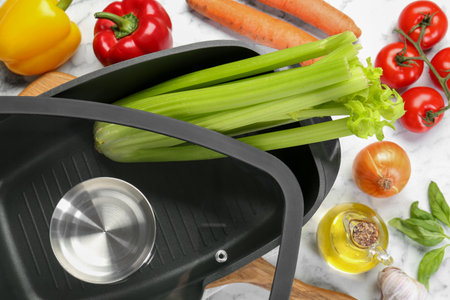 Black pot with glass lid, spices and vegetables on white marble table, flat lay